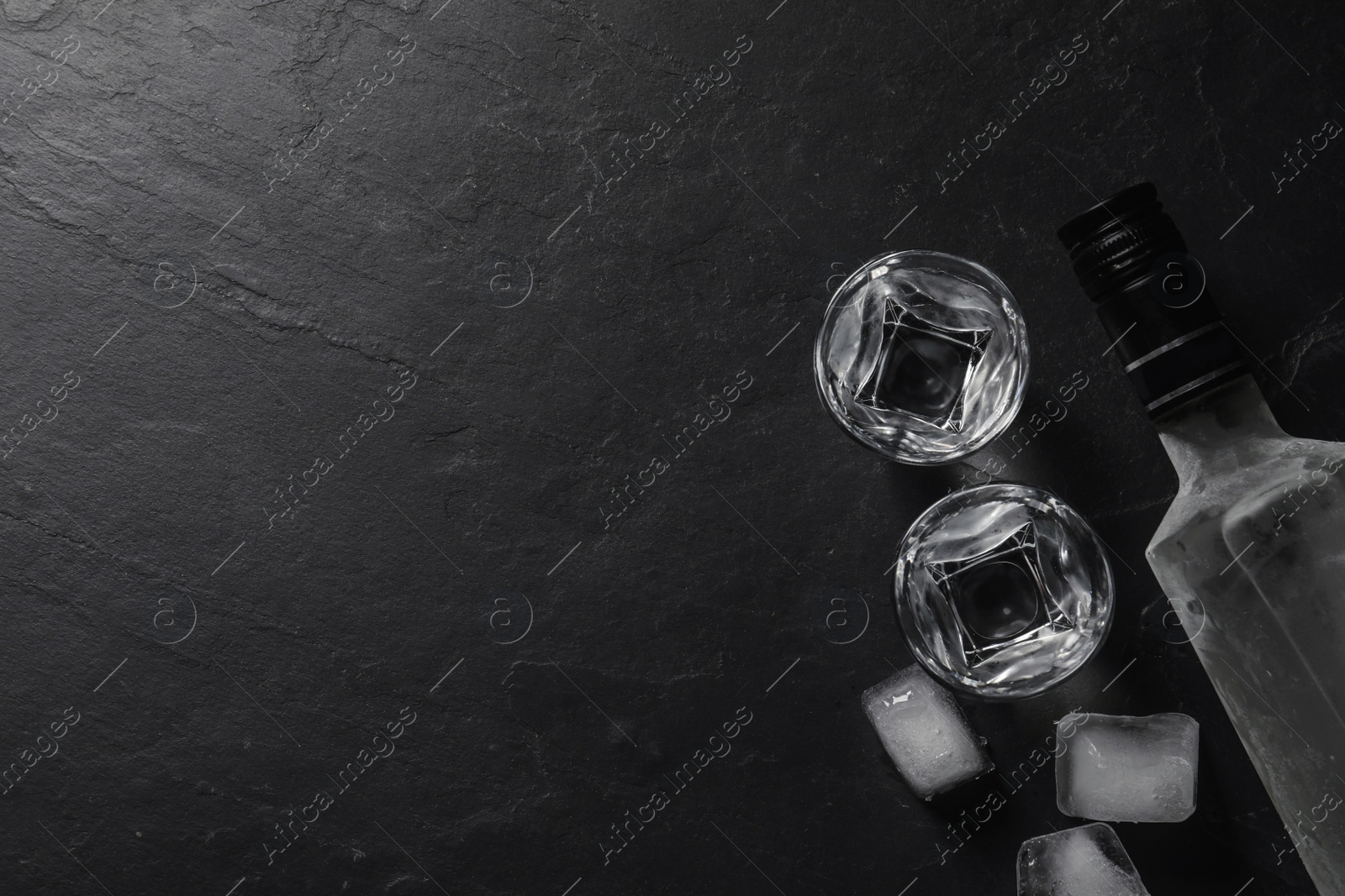 Photo of Bottle of vodka, shot glasses and ice cubes on black table, flat lay. Space for text