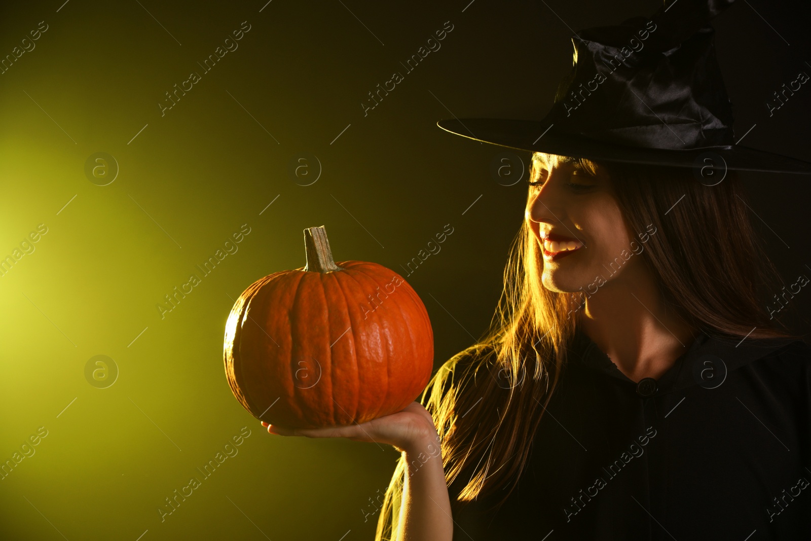 Photo of Young woman wearing witch costume with pumpkin on dark background. Halloween party