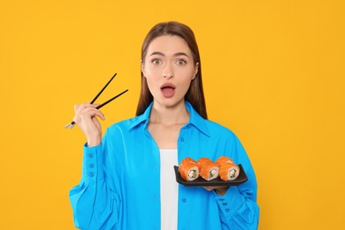 Emotional young woman with plate of sushi rolls and chopsticks on orange background