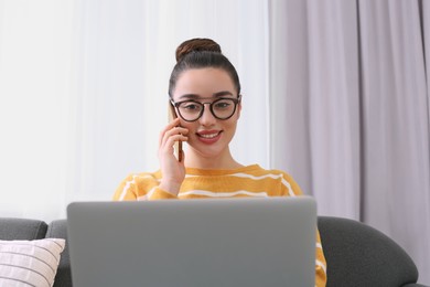 Home workplace. Happy woman with laptop talking on smartphone on sofa in room