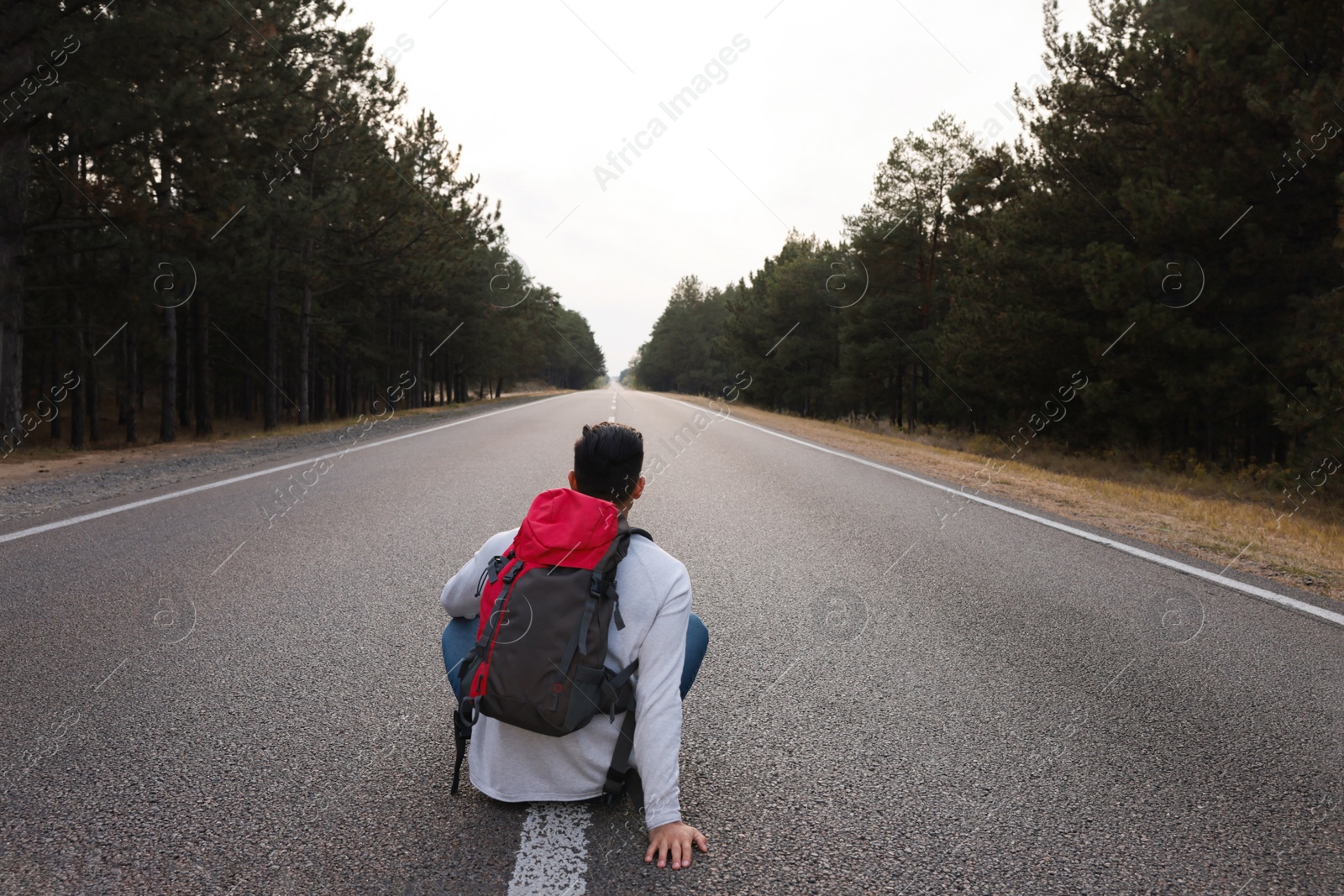 Photo of Man with backpack sitting on road near forest, back view