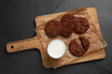 Photo of Board with tasty chocolate cookies and glass of milk on dark table, top view