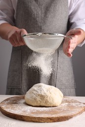 Man sprinkling flour over dough at table near grey wall, closeup