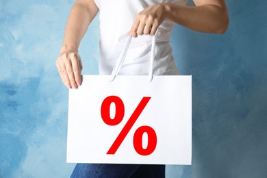 Image of Woman holding paper shopping bag with percent sign near light blue wall, closeup. Discount concept