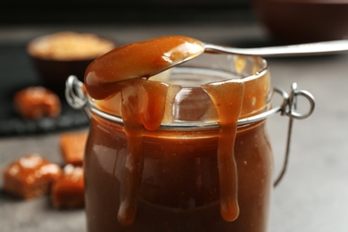 Photo of Jar with tasty caramel sauce and spoon on table, closeup