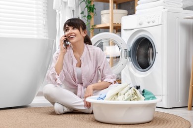 Photo of Happy young housewife with laundry talking on smartphone near washing machine at home
