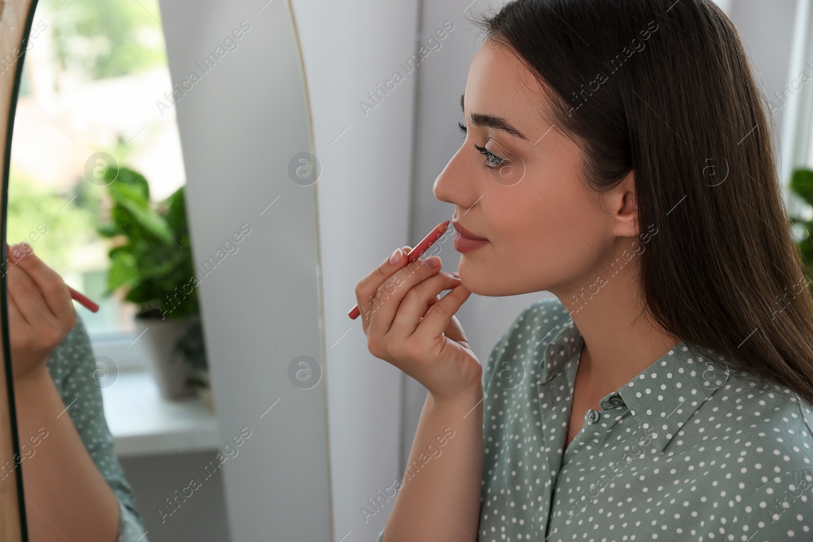 Photo of Beautiful young woman applying cosmetic pencil on lips near mirror indoors