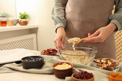 Photo of Making granola. Woman putting oat flakes into bowl at table in kitchen, closeup