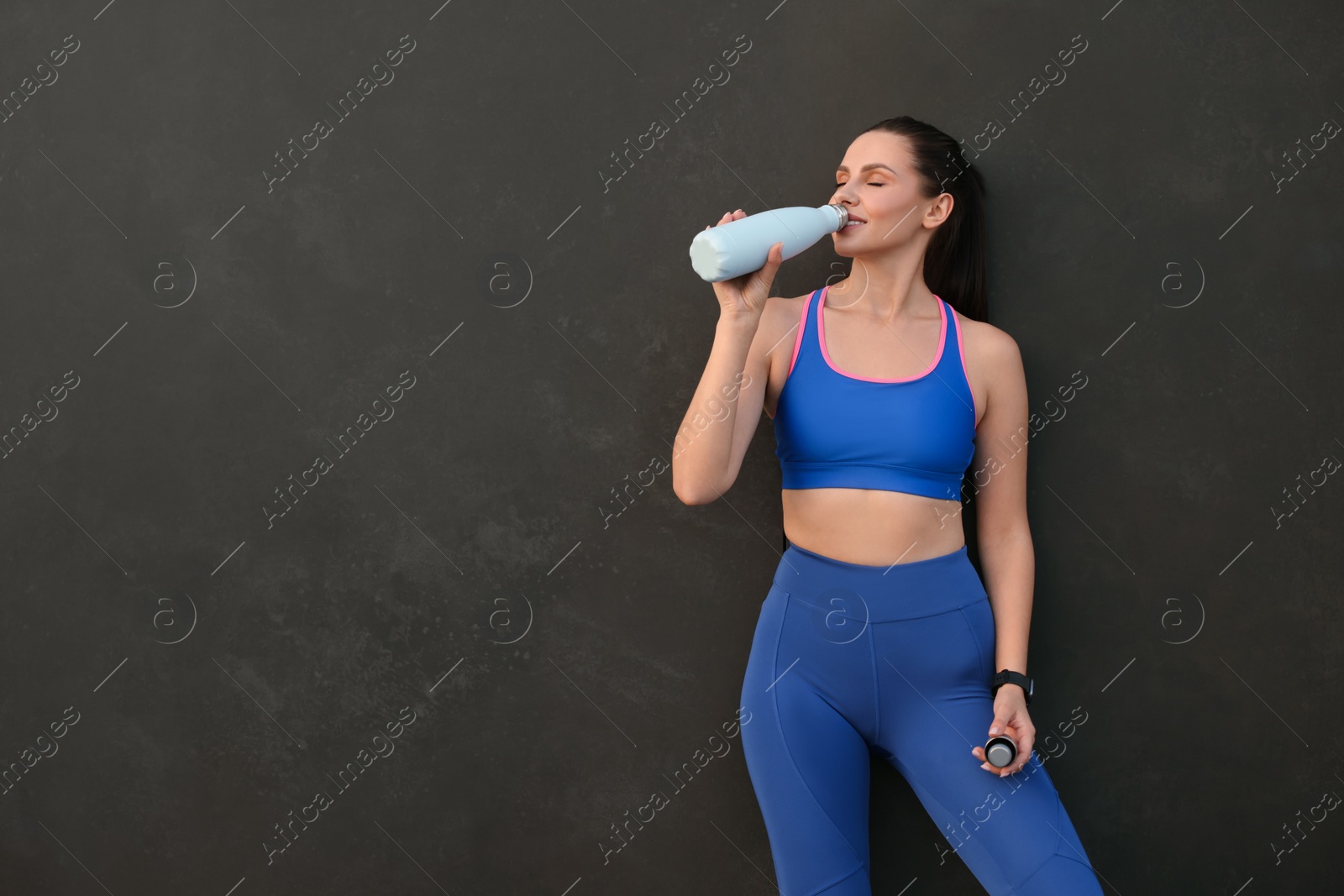 Photo of Beautiful woman in stylish sportswear drinking water near black wall, space for text