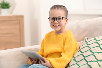 Photo of Portrait of cute little girl in glasses with tablet on sofa indoors