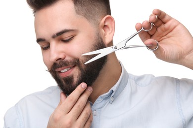 Photo of Handsome young man trimming beard with scissors on white background