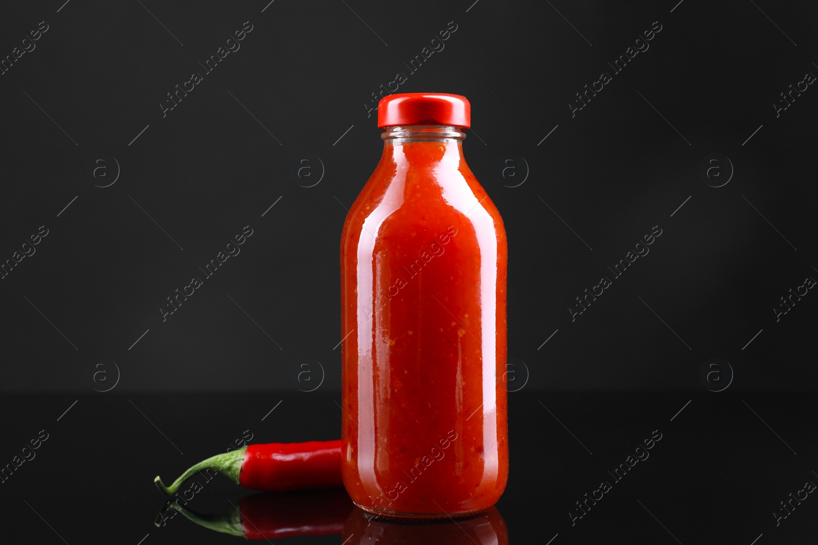 Photo of Spicy chili sauce in bottle and pepper against dark background