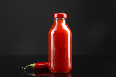 Photo of Spicy chili sauce in bottle and pepper against dark background