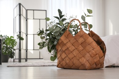 Stylish wicker basket with fresh eucalyptus branches and flowers on floor in bedroom