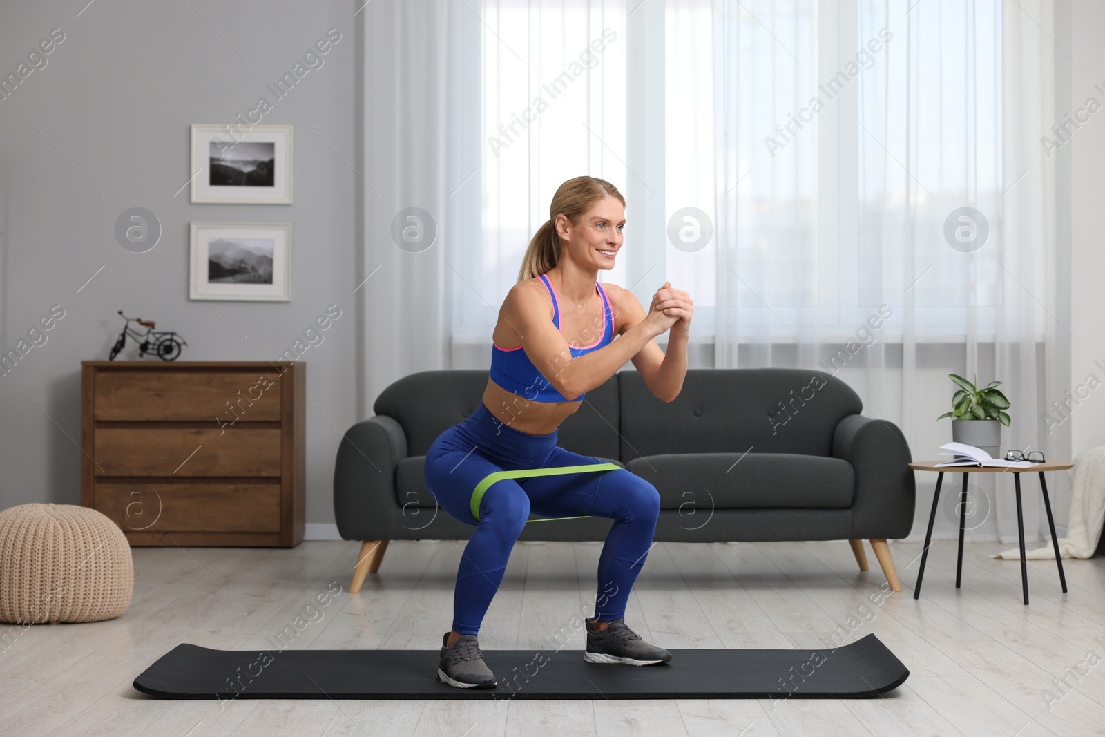 Photo of Athletic woman doing squats with fitness elastic band on mat at home