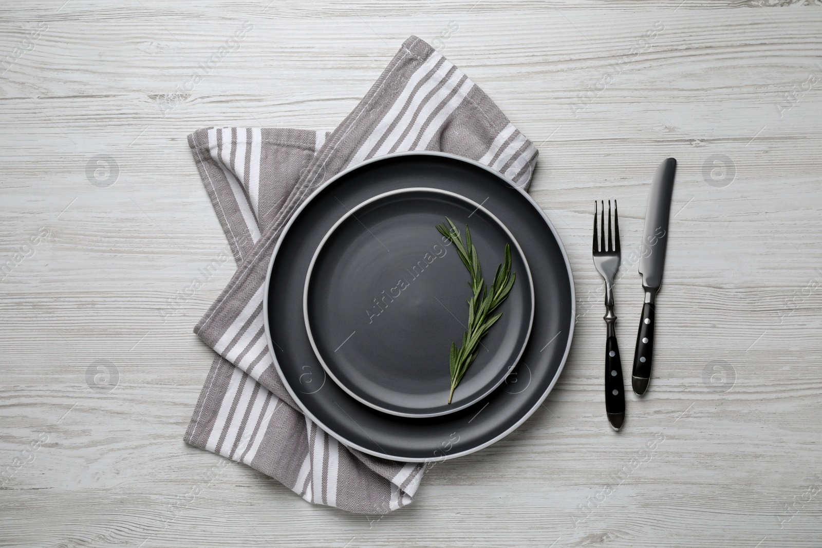 Photo of New dark plates with rosemary and cutlery on light wooden table, flat lay