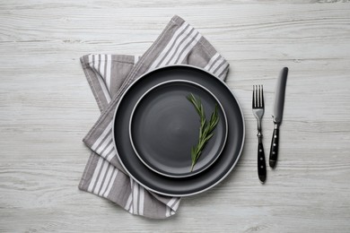 Photo of New dark plates with rosemary and cutlery on light wooden table, flat lay