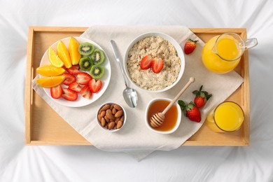 Photo of Tasty breakfast served in bed. Oatmeal, juice, fruits, almonds and honey on tray, top view