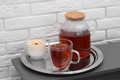 Teapot, cup of aromatic tea and burning candle on grey table near white brick wall