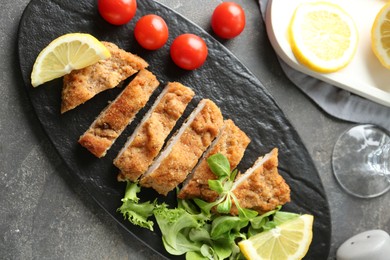 Photo of Tasty cut schnitzel served with tomatoes, greens and lemon on grey table, flat lay