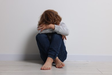 Photo of Child abuse. Upset boy sitting on floor near white wall