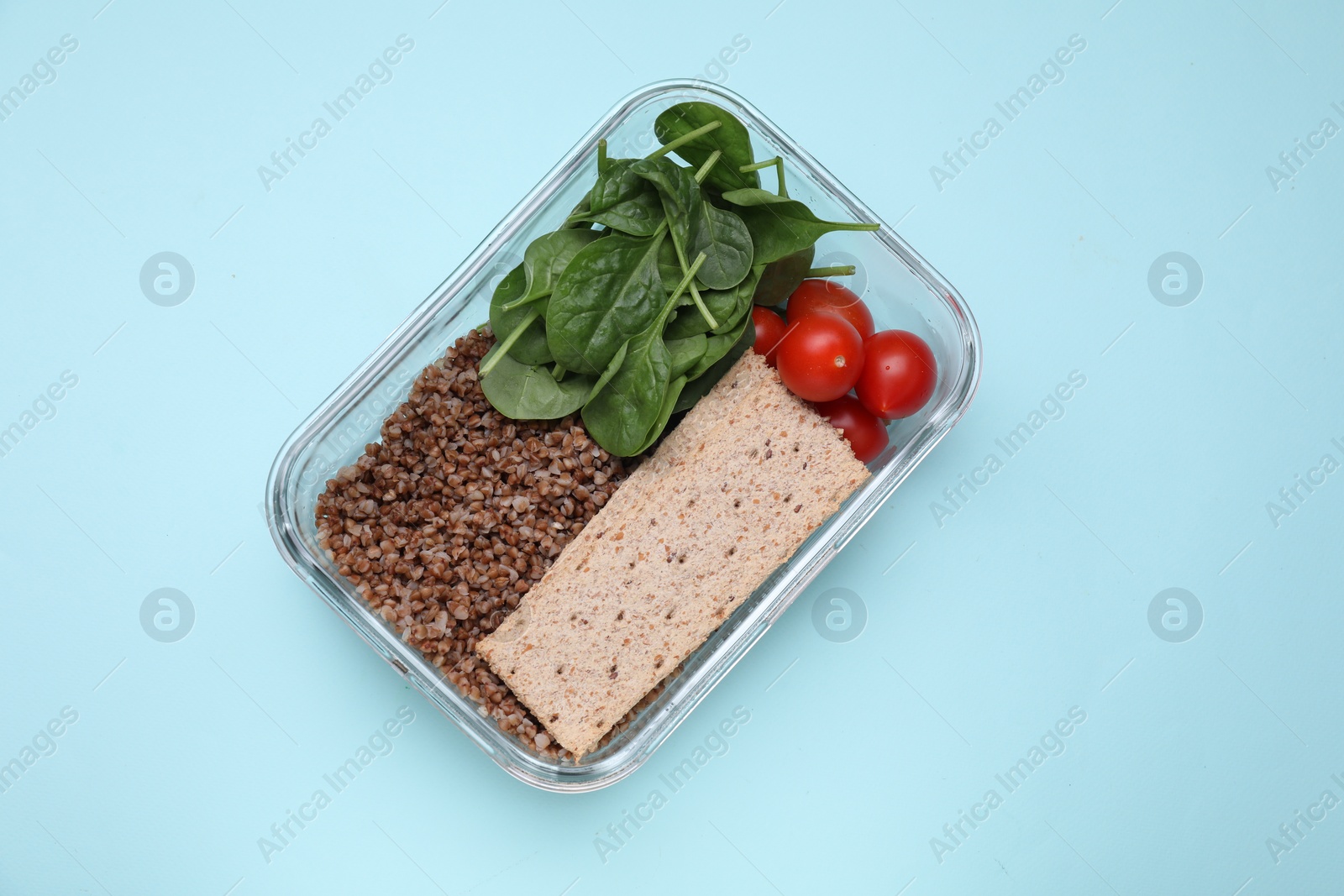 Photo of Glass container with buckwheat, fresh tomato, spinach and crispbreads on light blue background, top view