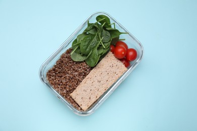 Photo of Glass container with buckwheat, fresh tomato, spinach and crispbreads on light blue background, top view