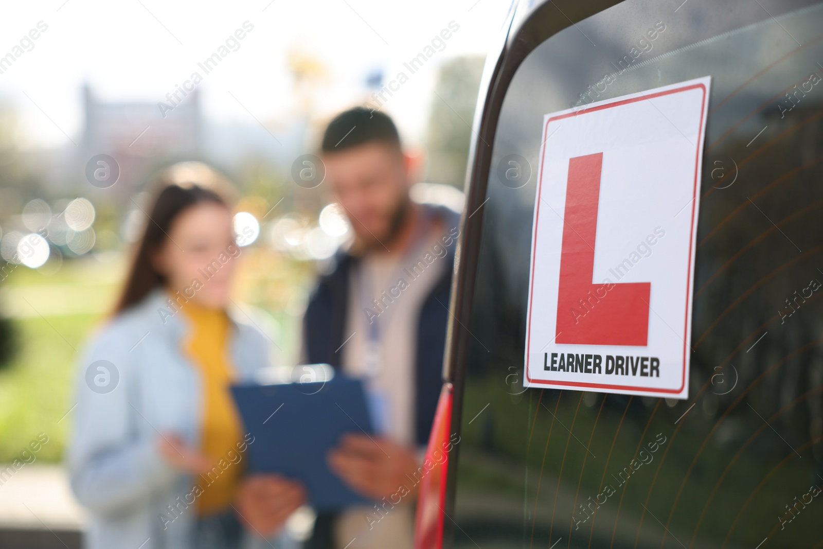 Photo of Learner driver and instructor with clipboard near car outdoors, selective focus on L-plate. Driving school