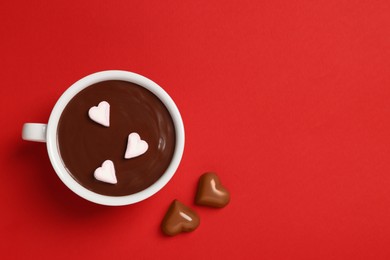 Cup of hot chocolate with heart shaped marshmallows and tasty candies on red background, flat lay. Space for text