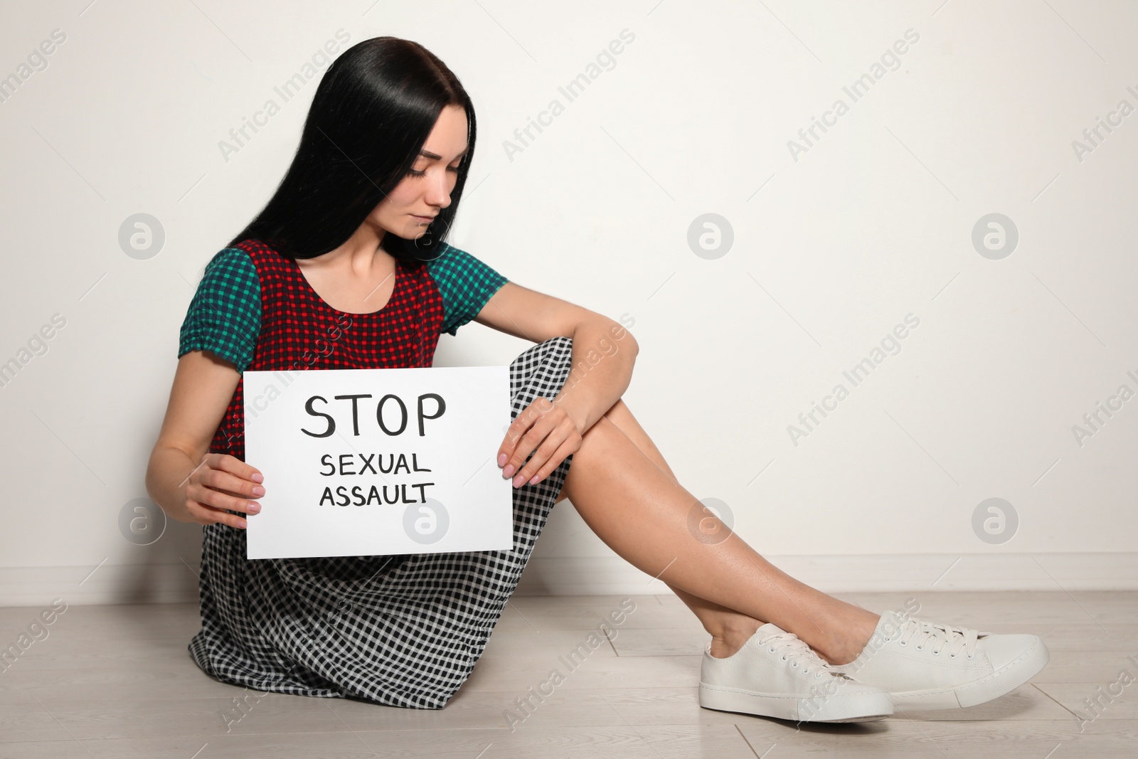 Photo of Young woman holding card with words STOP SEXUAL ASSAULT while sitting near light wall