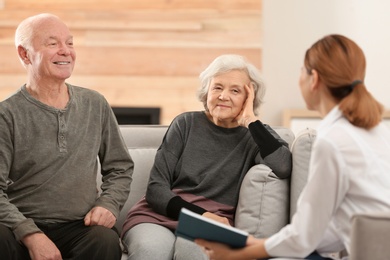 Photo of Female caregiver reading book to elderly spouses in living room