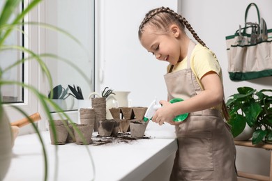 Little girl spraying water onto vegetable seeds in peat pots on window sill indoors