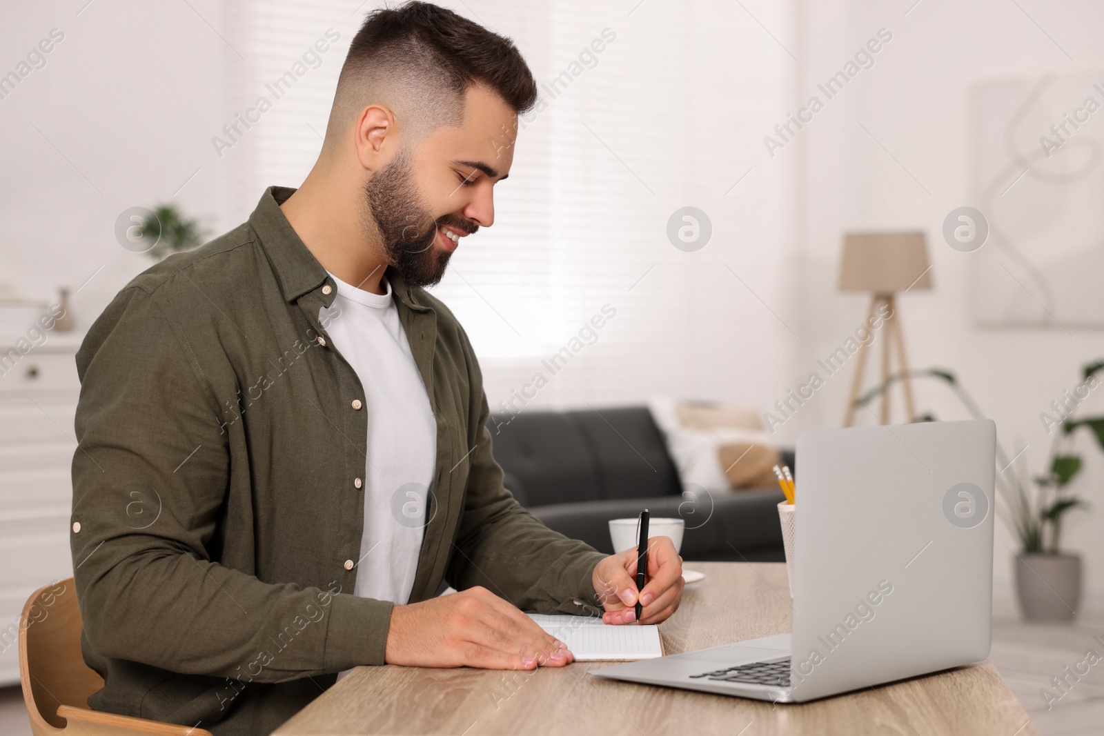 Photo of Young man writing in notebook at wooden table indoors