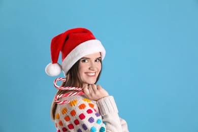 Photo of Pretty woman in Santa hat and festive sweater holding candy canes on light blue background, space for text