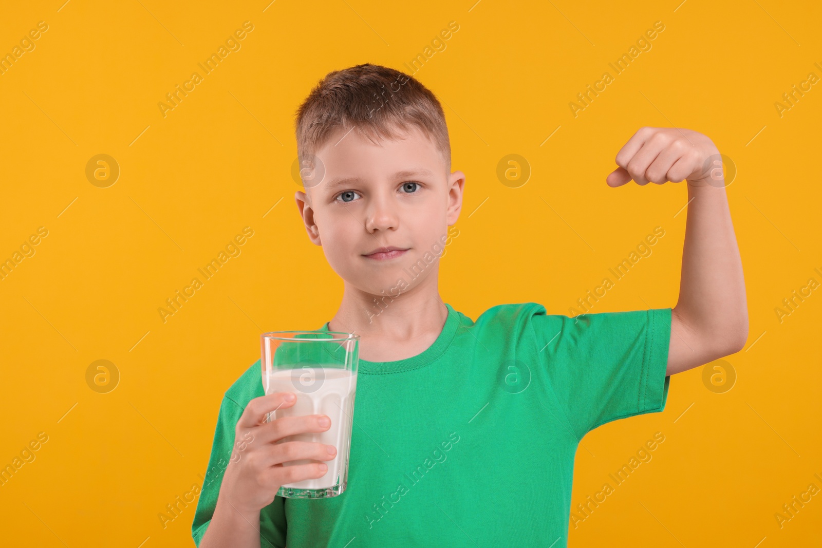 Photo of Cute boy with glass of fresh milk showing his strength on orange background