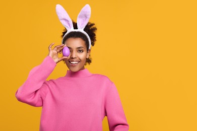 Happy African American woman in bunny ears headband covering eye with Easter egg on orange background, space for text