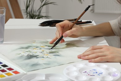 Woman painting flowers with watercolor at white table indoors, closeup. Creative artwork