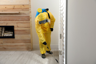 Photo of Pest control worker in protective suit spraying insecticide on floor at home