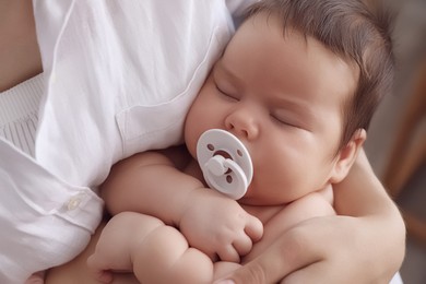 Photo of Mother holding her cute little baby with pacifier on blurred background, closeup