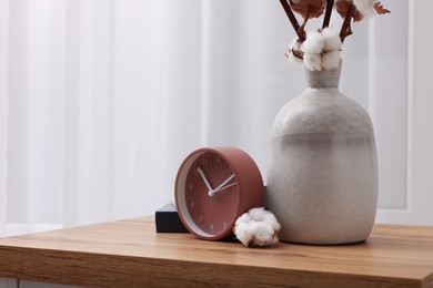 Photo of Alarm clock and vase with cotton branches on wooden table indoors. Space for text