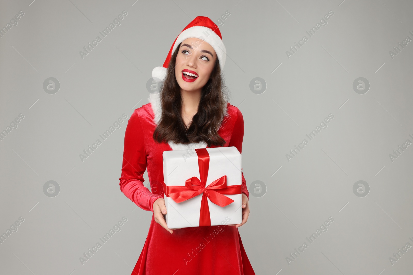 Photo of Beautiful young woman in red dress and Santa hat with Christmas gift on grey background