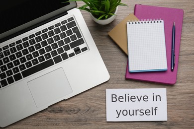 Photo of Note with motivational quote Believe in yourself, modern laptop and office stationery on wooden table, flat lay