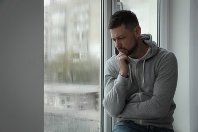 Photo of Depressed man near window at home. Space for text
