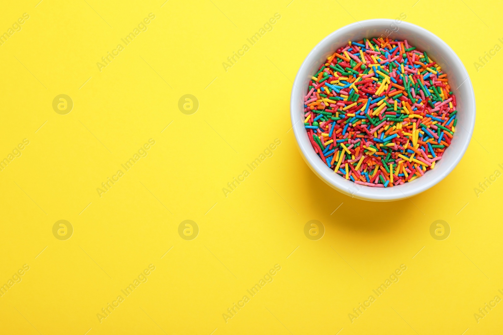 Photo of Colorful sprinkles in bowl on yellow background, top view with space for text. Confectionery decor