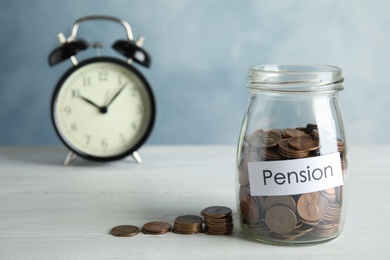 Glass jar with label PENSION and coins near alarm clock on white table