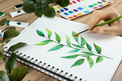 Woman painting green twig in sketchbook at wooden table, closeup