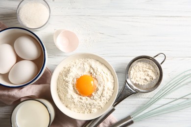 Photo of Flour with yolk in bowl and other ingredients for dough on white wooden table, flat lay