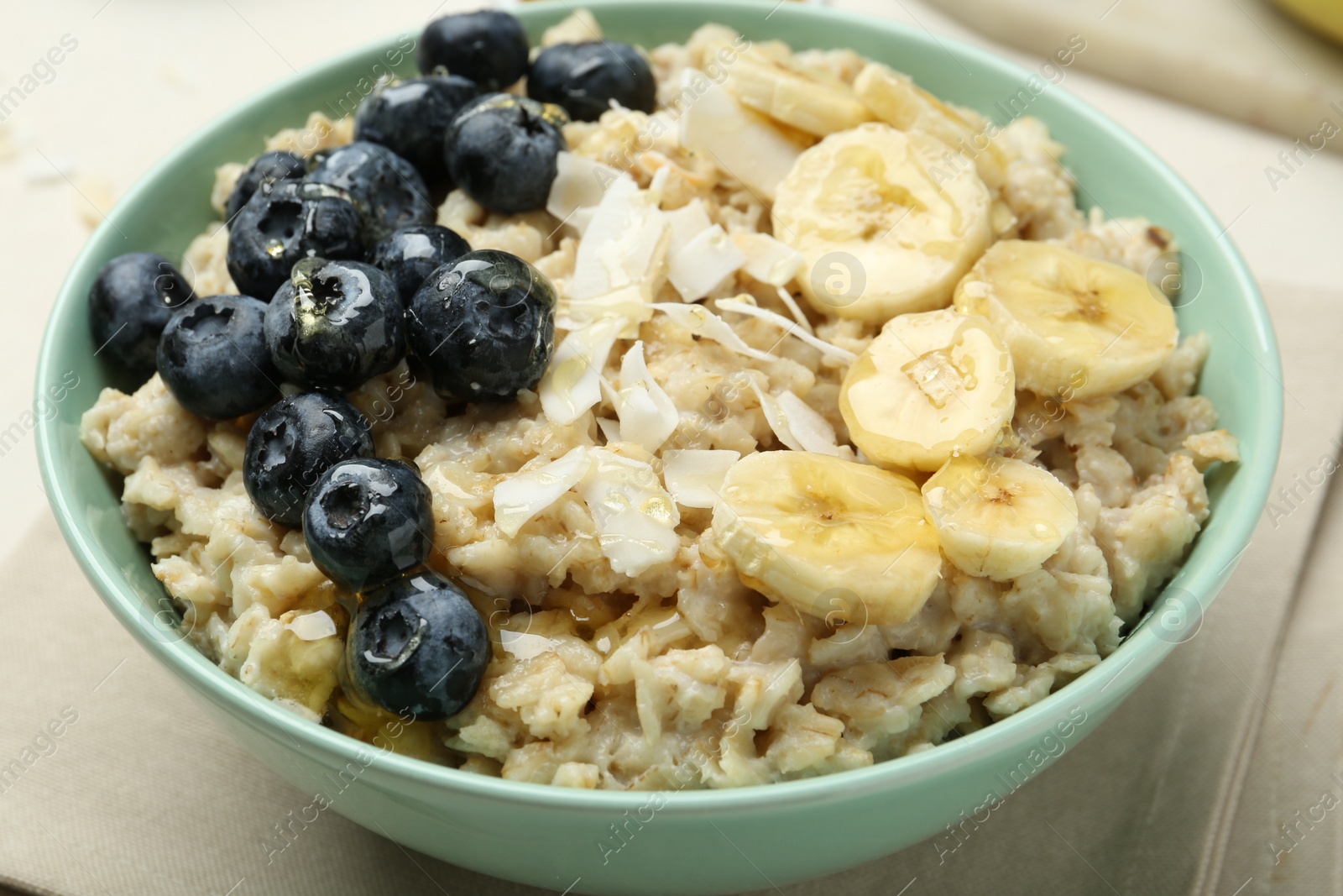 Photo of Tasty oatmeal with banana, blueberries, coconut flakes and honey served in bowl on beige table, closeup