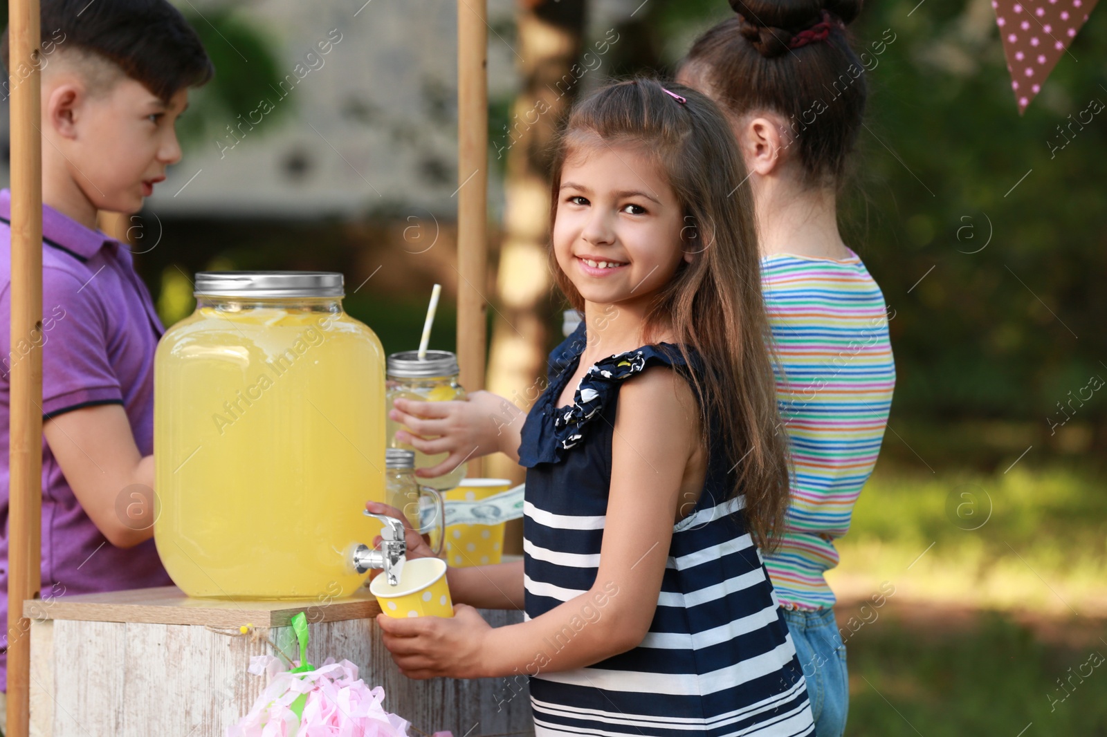 Photo of Cute little girl pouring natural lemonade into cup in park. Summer refreshing drink