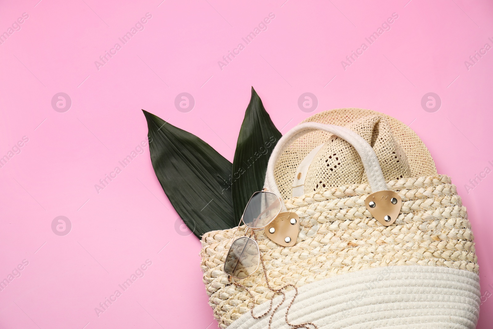 Photo of Elegant woman's straw bag with hat, tropical leaves and sunglasses on pink background, top view